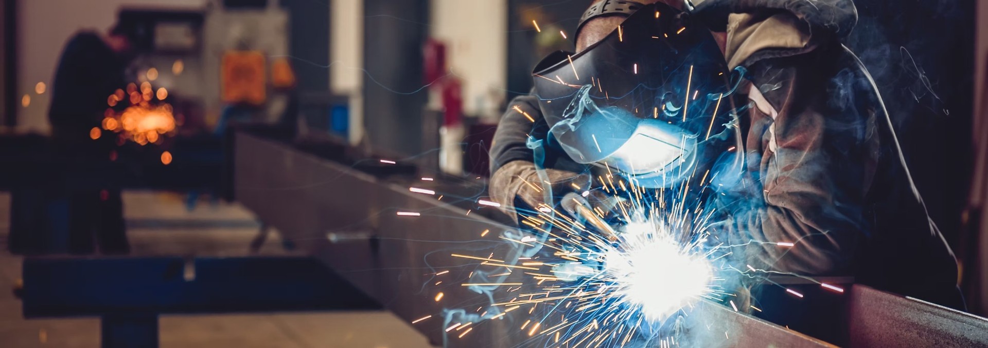 A welder working on a metal structure, with another worker in the background.
